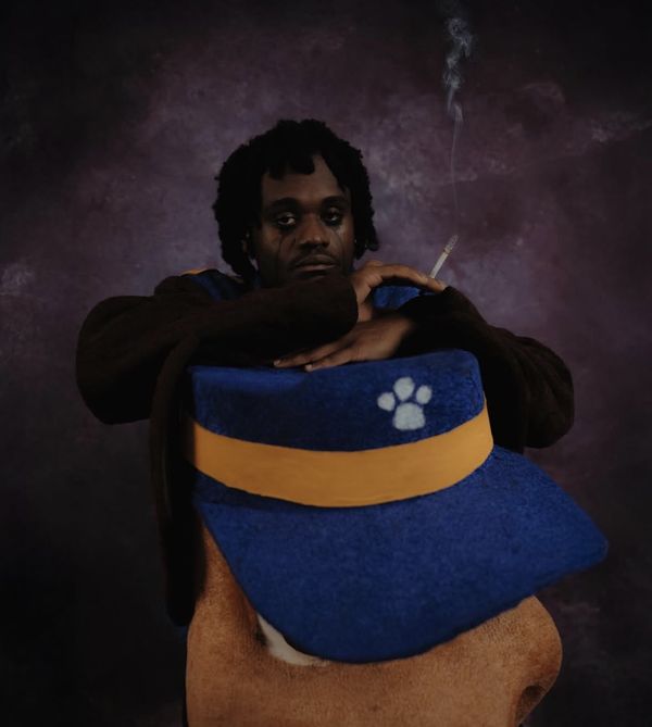 Portrait with dark tear-streak makeup, arms folded over an oversized plush blue-and-gold cap, smoke rising against a purple backdrop.