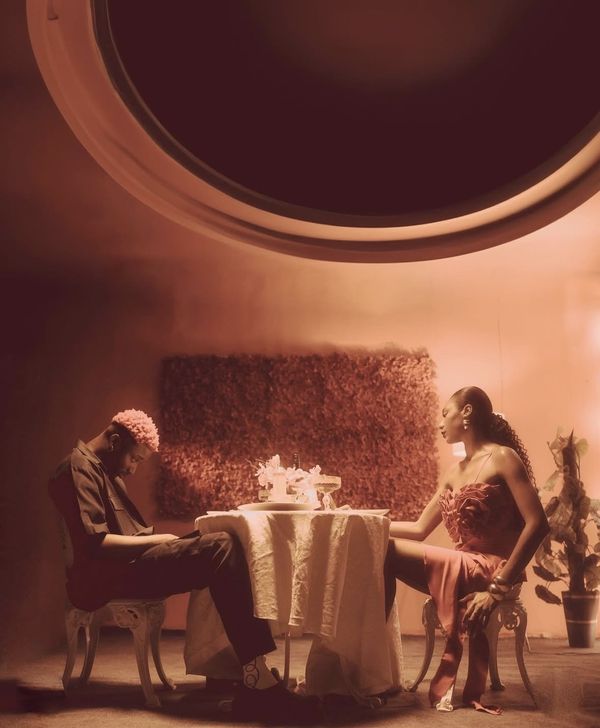 A couple at a candlelit table under a huge circular window, the man slumped, the woman watching him, in warm rose tones.