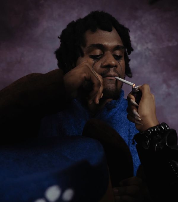 A hand with long nails lights a cigarette for a man in a blue fleece, eyes lowered.