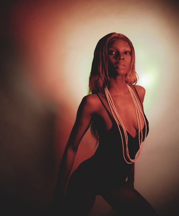 A woman in a black bodysuit and long pearl strands, lit in red with a soft green glow behind.