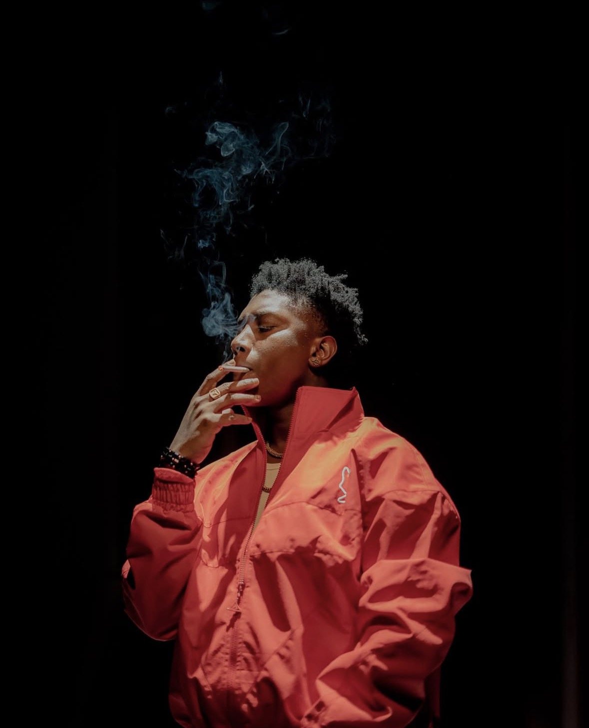 Portrait in a red jacket, exhaling smoke under a single hard light against black.