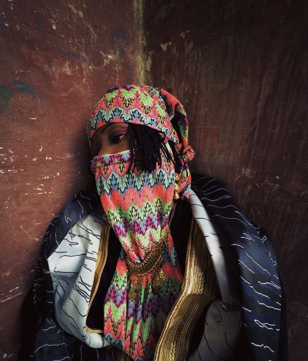 A woman veiled in bright zigzag-patterned fabric and gold coins leans into the corner of a weathered red wall.